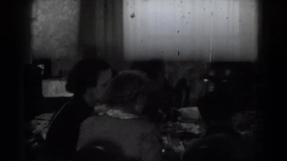 In a 1947 Lincoln, Nebraska home-movie frame, adults sit around a dim dining table near a bright window.