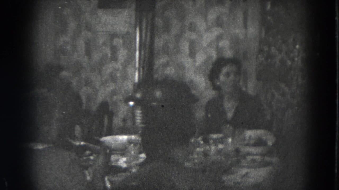 A woman and another adult sit at a crowded dinner table in the dim 1947 home-movie footage.