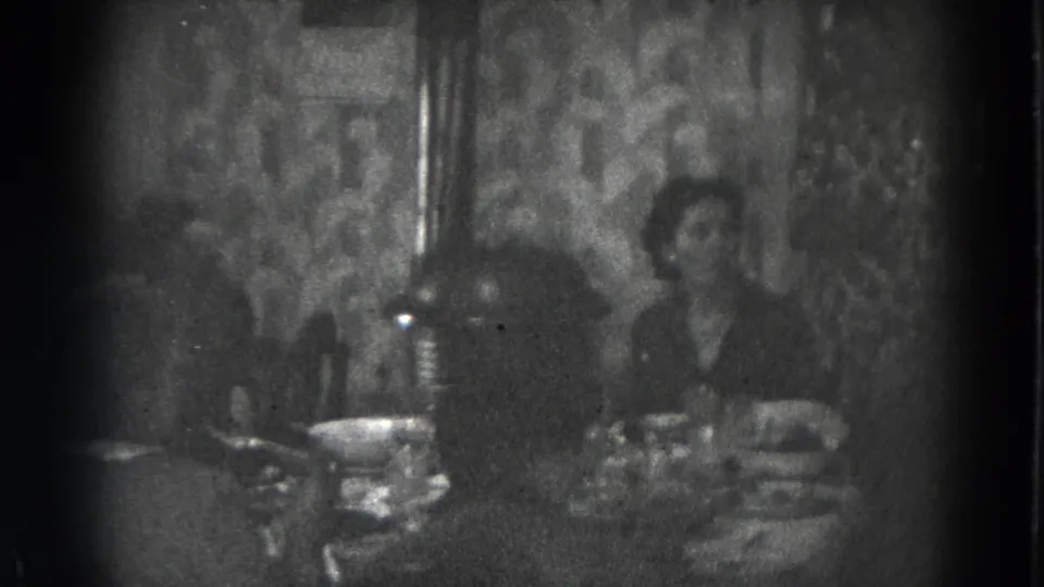 A woman and another adult sit at a crowded dinner table in the dim 1947 home-movie footage.