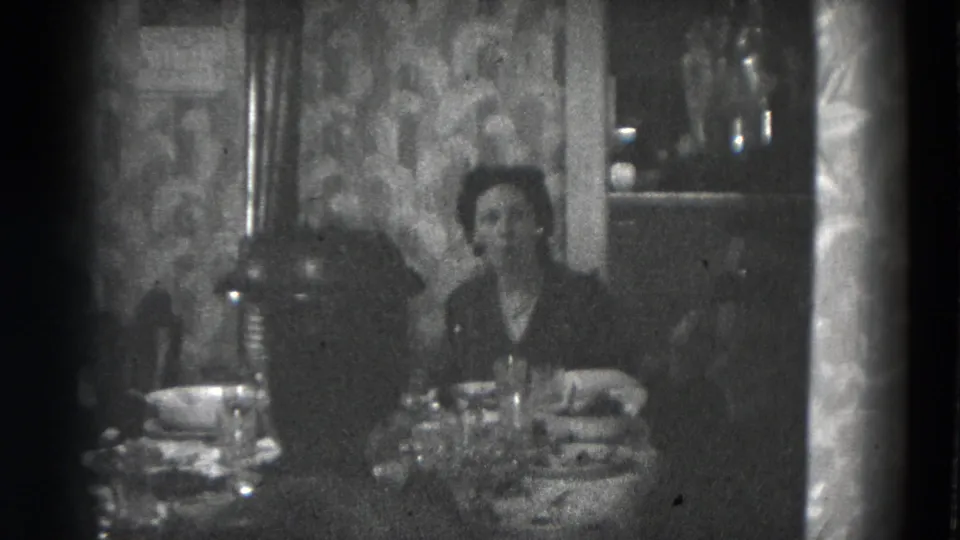 A woman faces the camera from a dining table in a private Lincoln home, with dishes and a lamp visible.