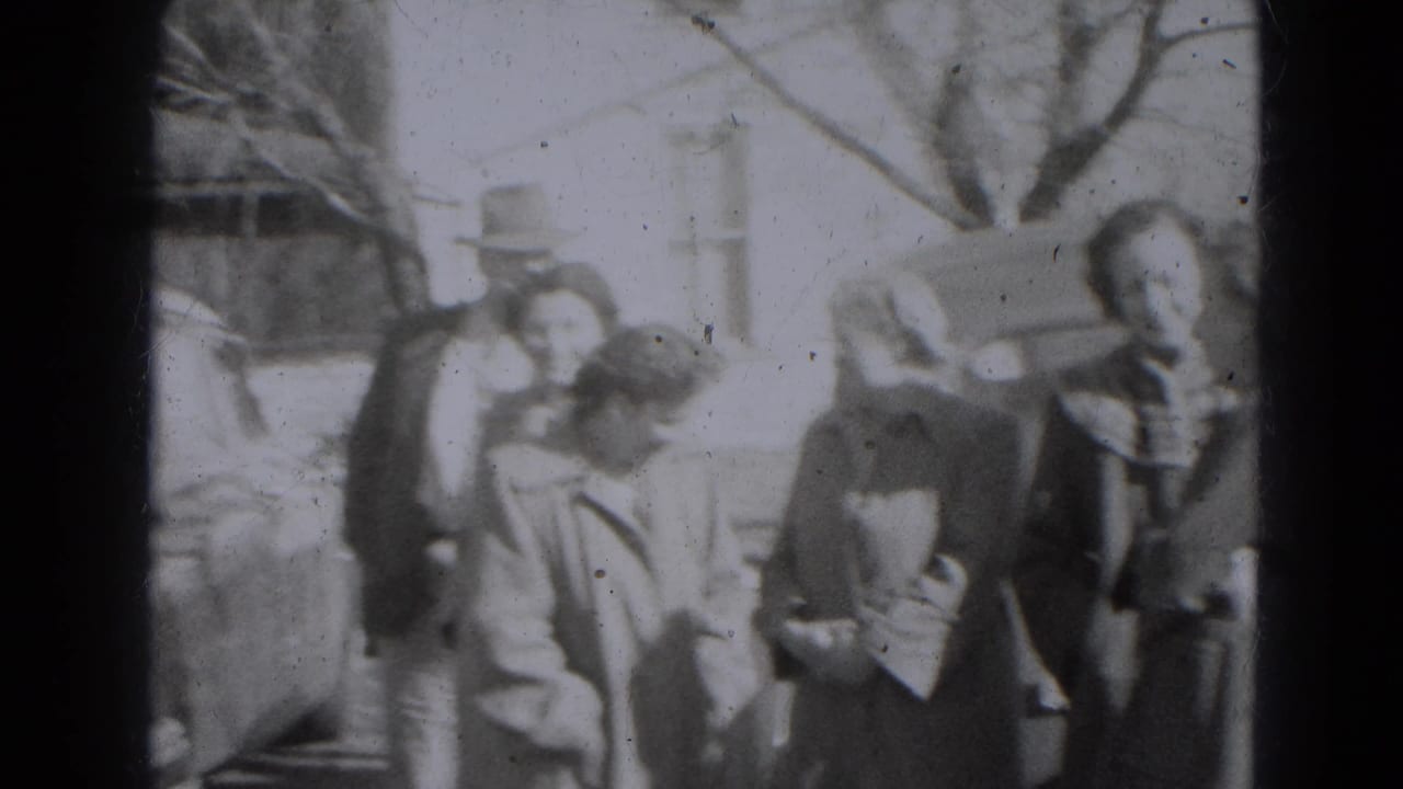 A group of adults in period outerwear stands and bends near a parked car in 1947 Lincoln, Nebraska.