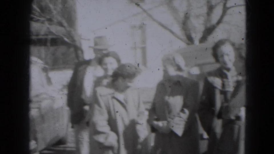 A group of adults in period outerwear stands and bends near a parked car in 1947 Lincoln, Nebraska.