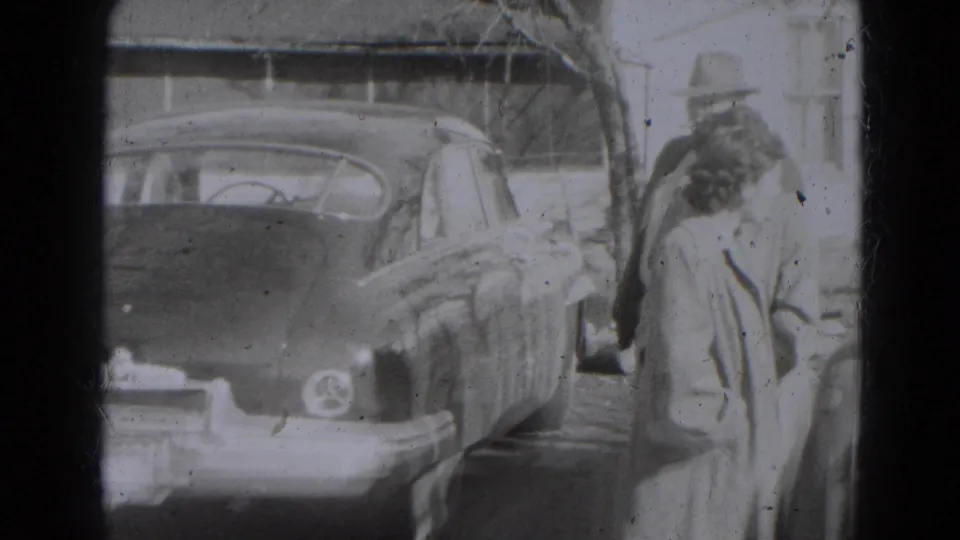 A woman in a long coat walks beside the rear of a dark car while a man in a hat stands nearby in Lincoln, Nebraska.