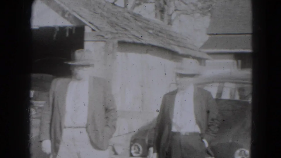 1947 Lincoln, Nebraska home-movie frame showing adults in period coats and hats gathered by parked cars outside a building.