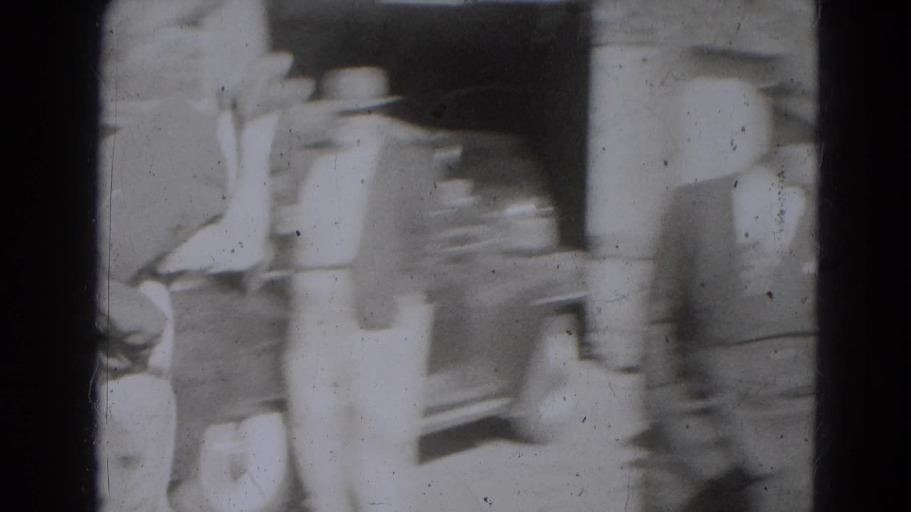 A blurred adult in period clothing moves past a building entrance, with a parked car and other figures partly visible nearby.