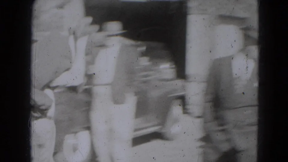 A blurred adult in period clothing moves past a building entrance, with a parked car and other figures partly visible nearby.