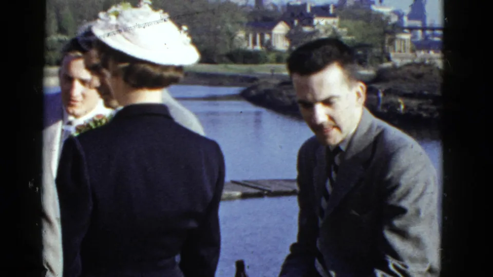 1951 Boston waterfront home-movie frame showing formally dressed adults conversing near water and distant buildings.