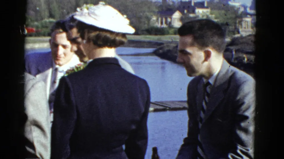 1951 Boston waterfront frame with a woman in a white hat and suited men standing in conversation near the water.