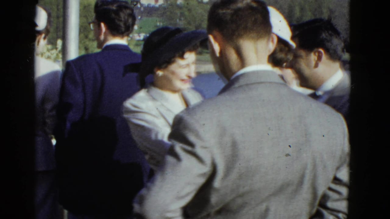A woman in a dark hat smiles while standing among men in suits outdoors.