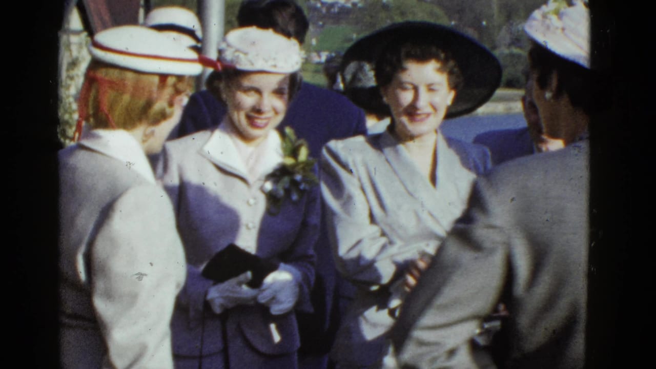 Three women in dress suits and hats smiling in a group outdoors.