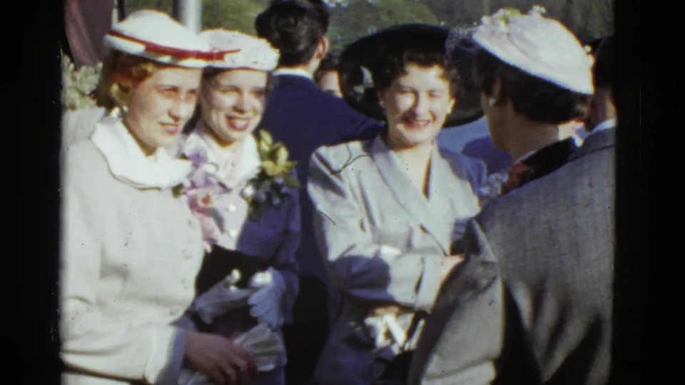 1951 Boston home-movie frame showing women in formal hats and suits talking in a crowded outdoor group.