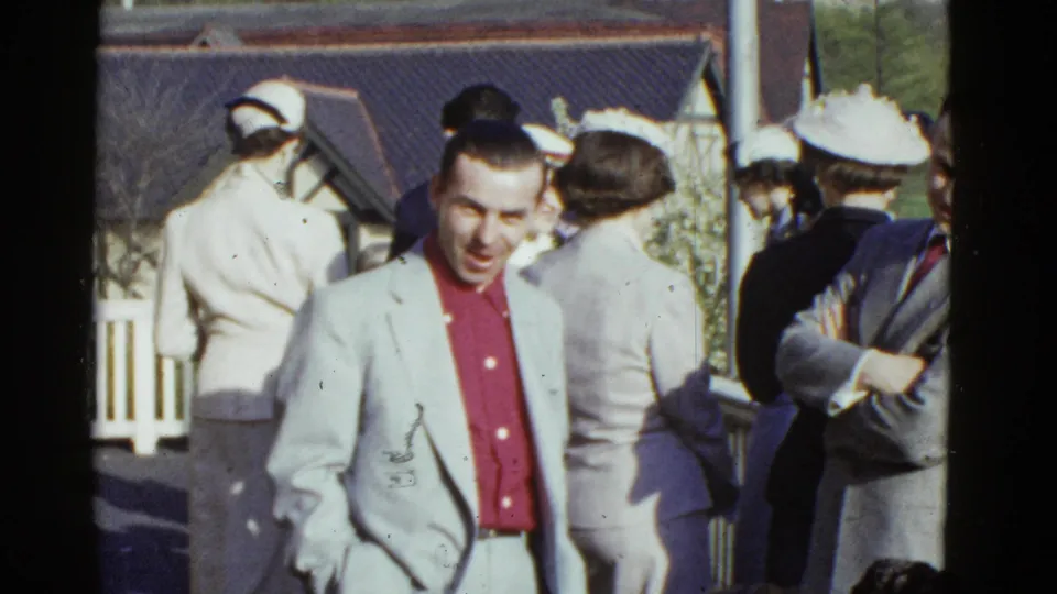 A man in a light jacket and red shirt stands among adults in formal clothing at the outdoor Boston social scene.