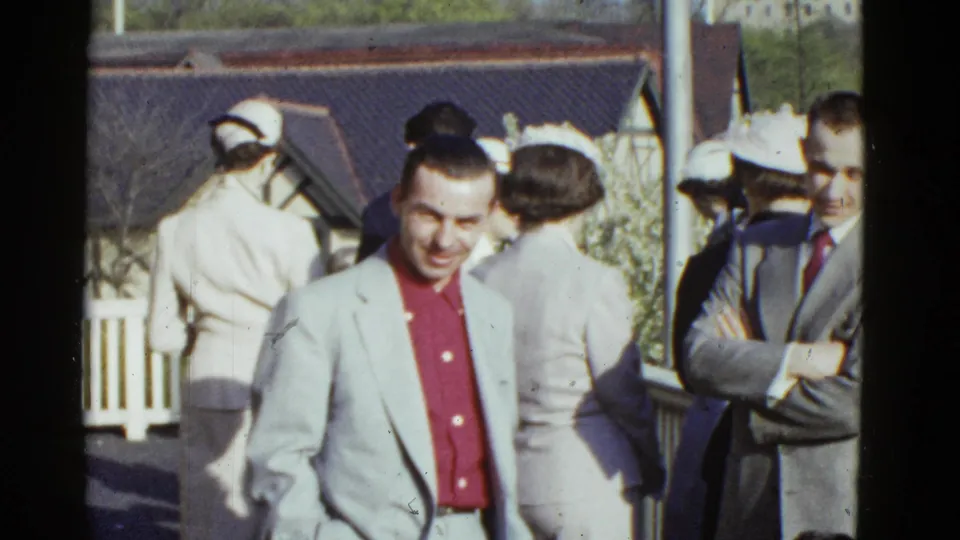 1951 Boston home-movie frame showing adults in suits and hats outdoors, with a smiling man in a light jacket and red shirt near the foreground.