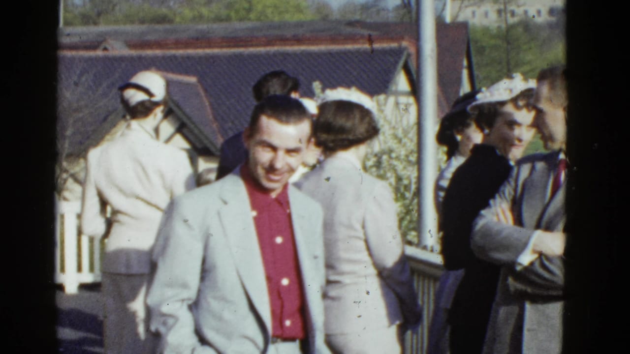 A man smiles at the camera while adults in hats and suits gather near a railing outdoors.
