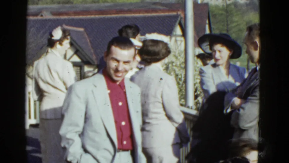 1951 Boston home-movie still of a sunny outdoor group scene, with a smiling man in front and women in hats nearby.