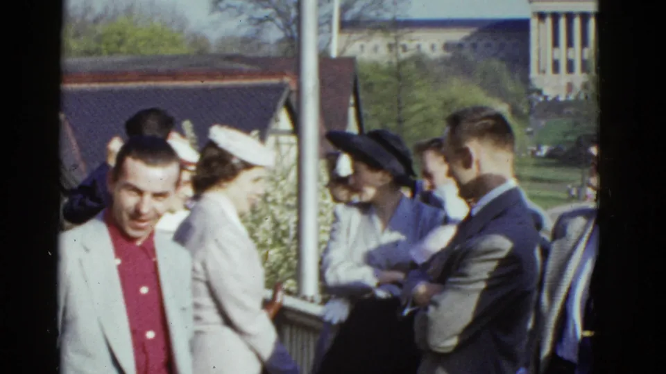 1951 Boston frame showing adults in mid-century formal daywear gathered outdoors, with trees, rooftops, and a railing behind them.