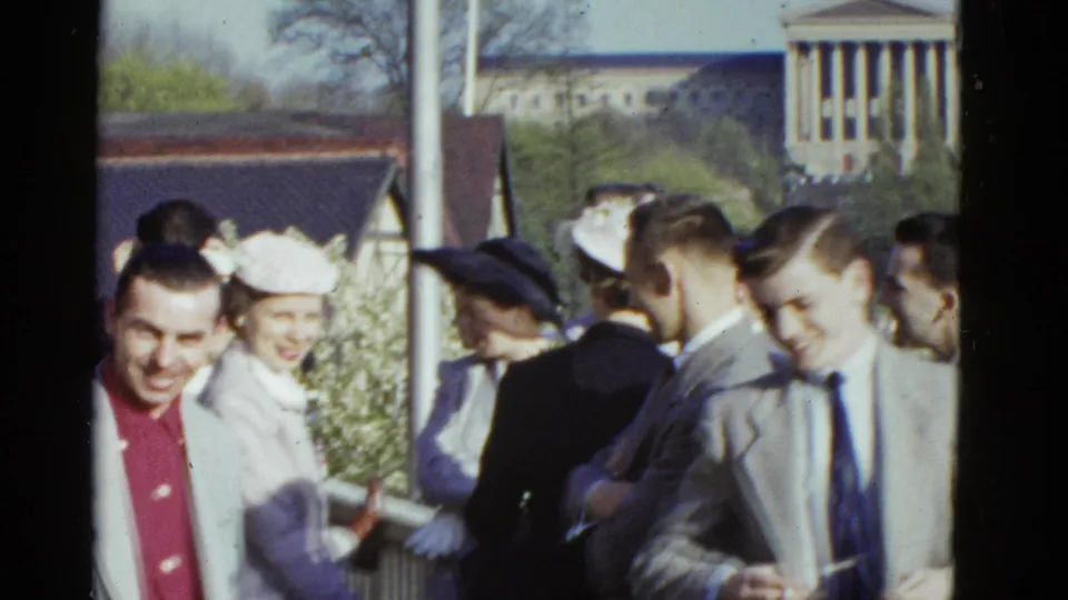 In 1951 Boston, young adults in suits, coats, gloves, and hats gather outdoors with a classical building behind them.