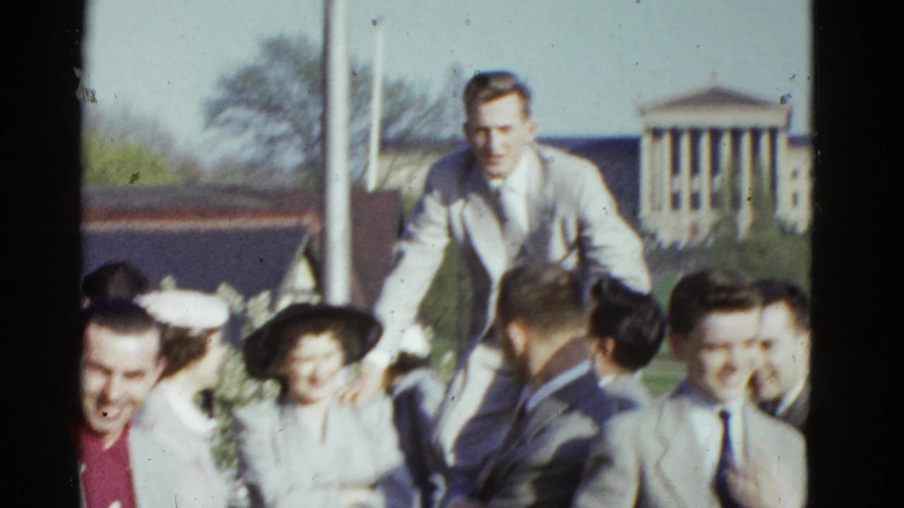 A man rises above a smiling group gathered outside a columned building.
