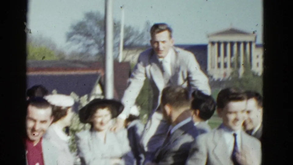 A formally dressed man stands or climbs above a group of smiling young adults outdoors in 1951 Boston.