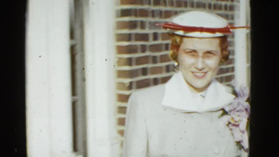 A woman in 1950s formal attire, including a light coat and white hat with trim, faces the camera near a brick wall in Boston.