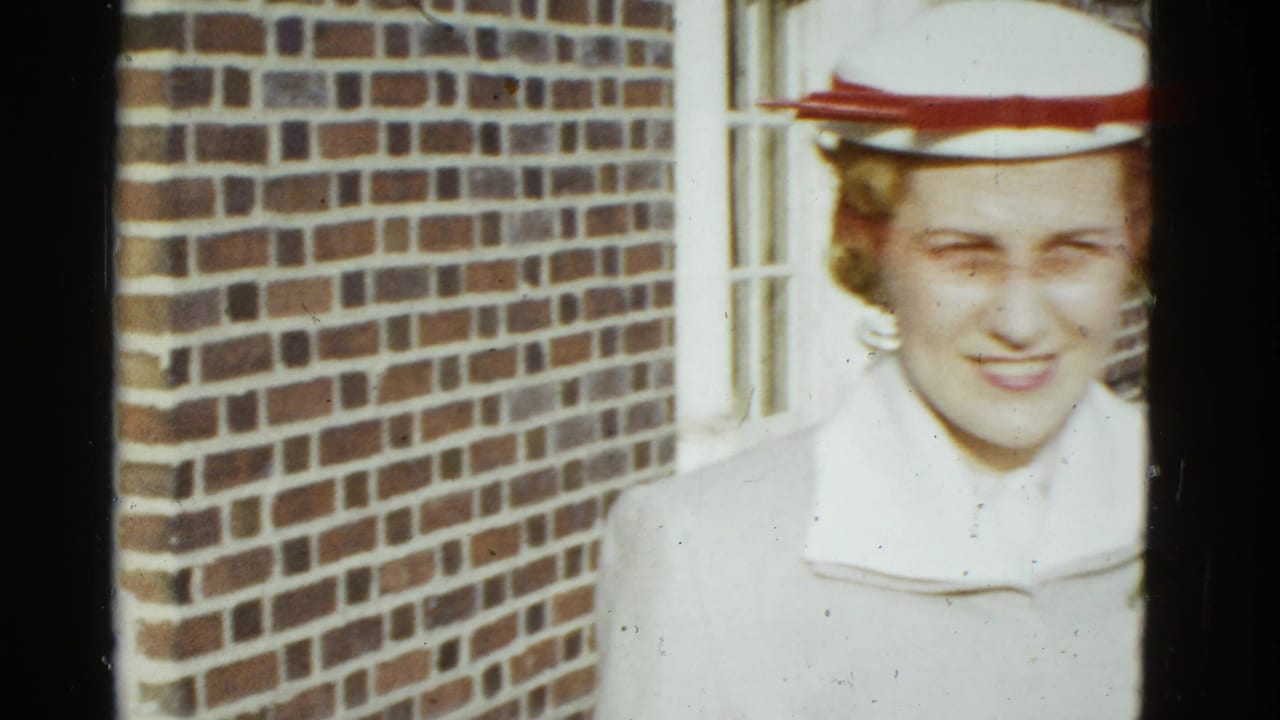 In 1951 Boston home-movie footage, a woman in a white coat and white hat with a red band stands by a brick wall and window.