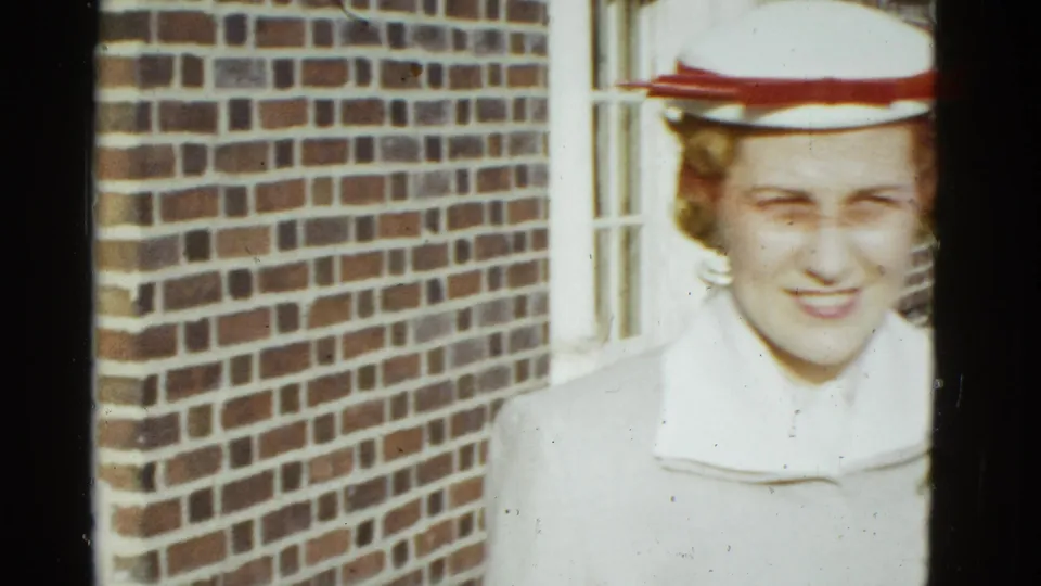 In 1951 Boston home-movie footage, a woman in a white coat and white hat with a red band stands by a brick wall and window.