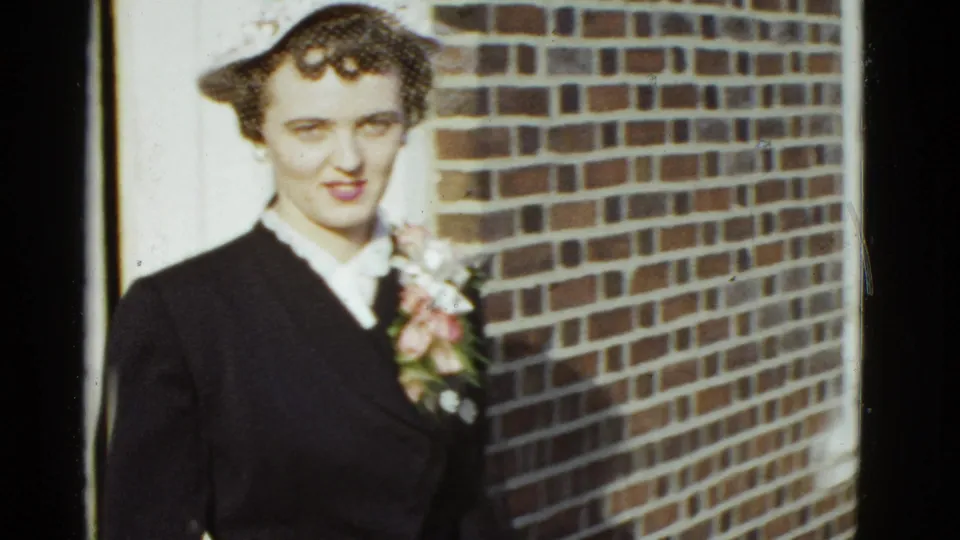 In 1951 Boston footage, a woman in a dark suit and small hat faces the camera with a floral corsage on her lapel.