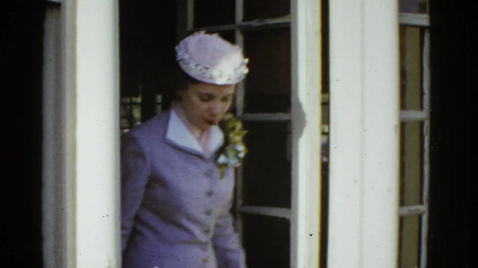 A woman in a lavender suit and matching hat walks by a doorway in 1951 Boston home-movie footage, with a corsage visible on her jacket.