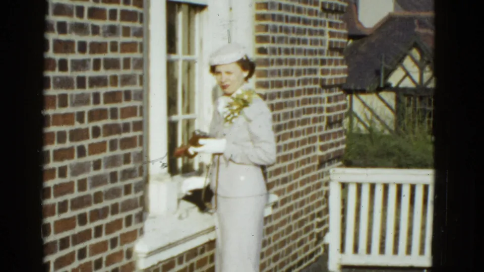 In 1951 Boston home-movie footage, a woman in a light suit and hat stands on a brick porch with flowers and a small camera.
