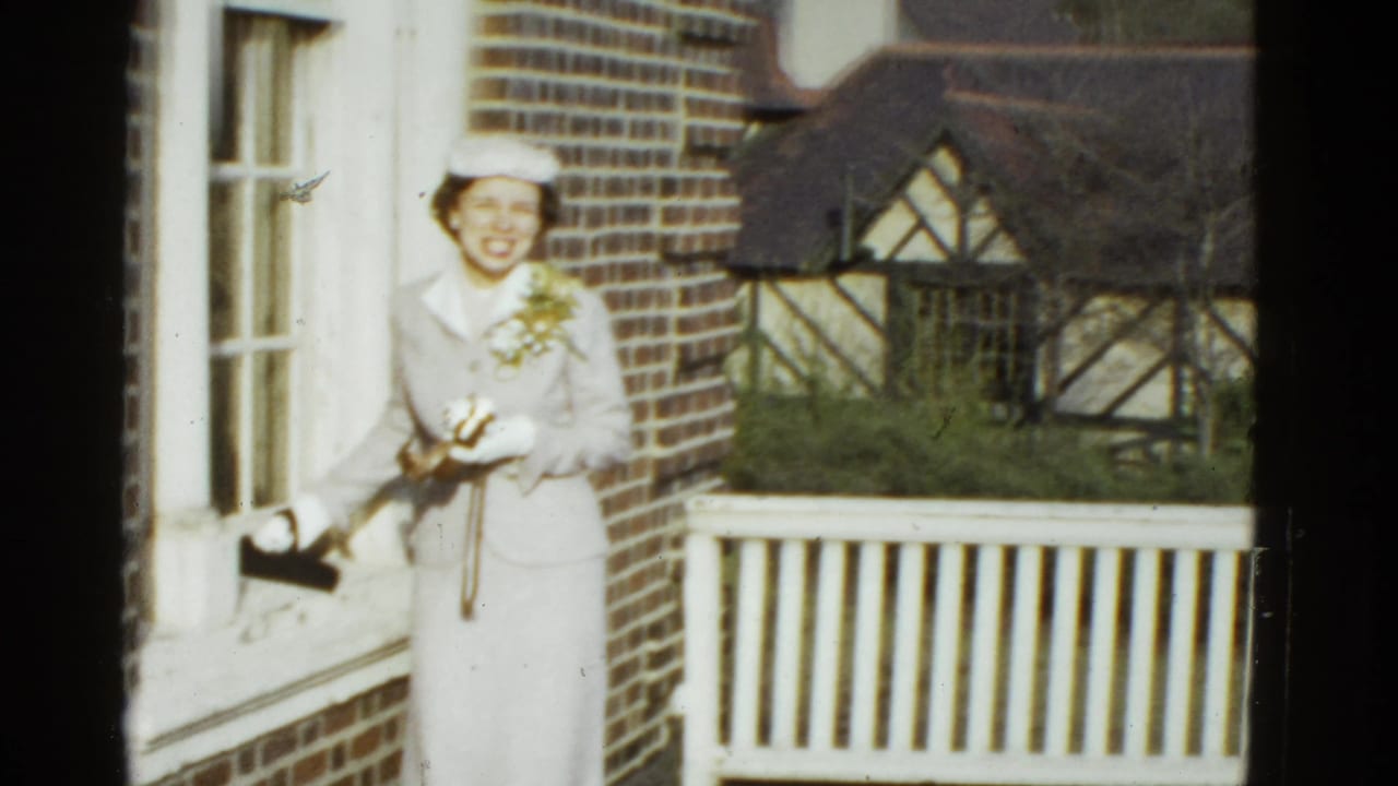 Smiling woman in a light suit and hat faces the camera on a brick porch with a white railing behind her.