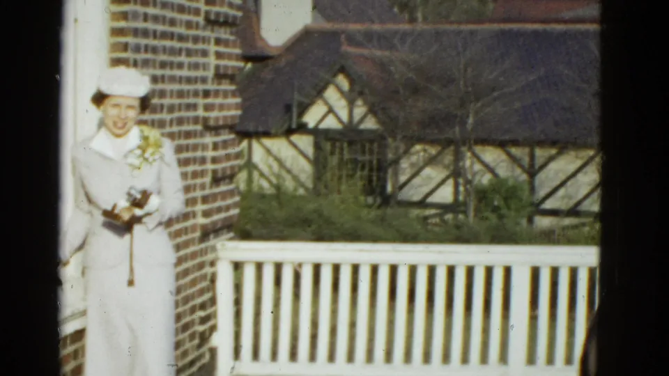 The woman remains posed on the Boston porch, framed by brickwork, white railing, and Tudor-style residential details in the background.