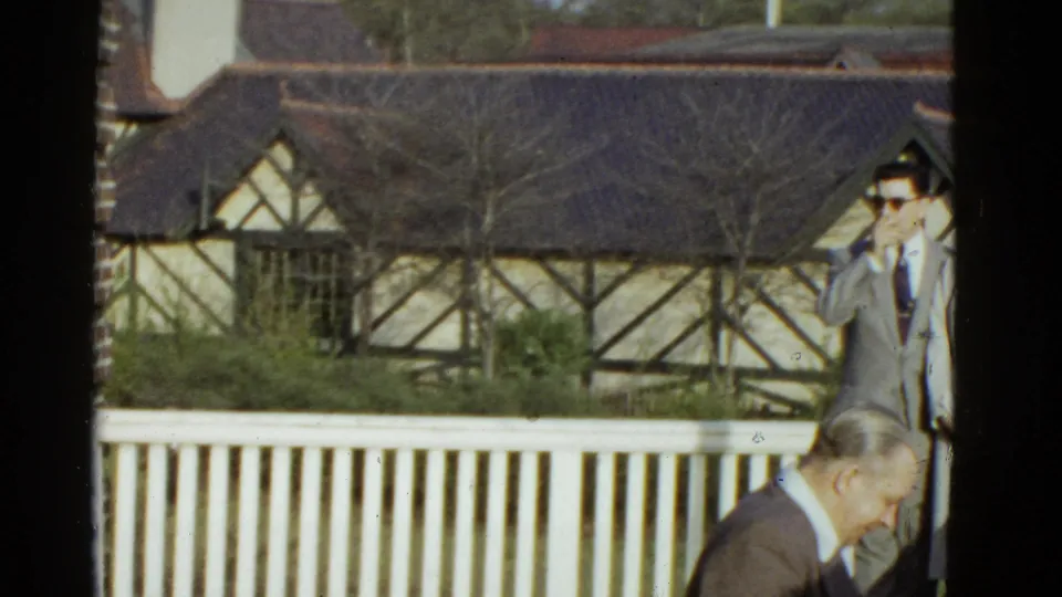 The camera shifts right from the porch scene, showing the white railing, Tudor-style building, and two adult men partly visible at the edge.