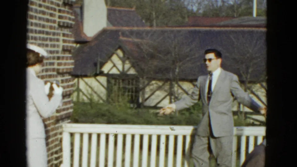 The man in a light suit stands with arms slightly out near the railing as a woman at left appears to photograph him in 1951 Boston footage.