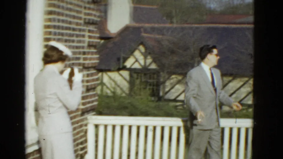 A woman in white faces a suited man near a brick wall and white railing, with a Tudor-style building behind them in the 1951 Boston clip.