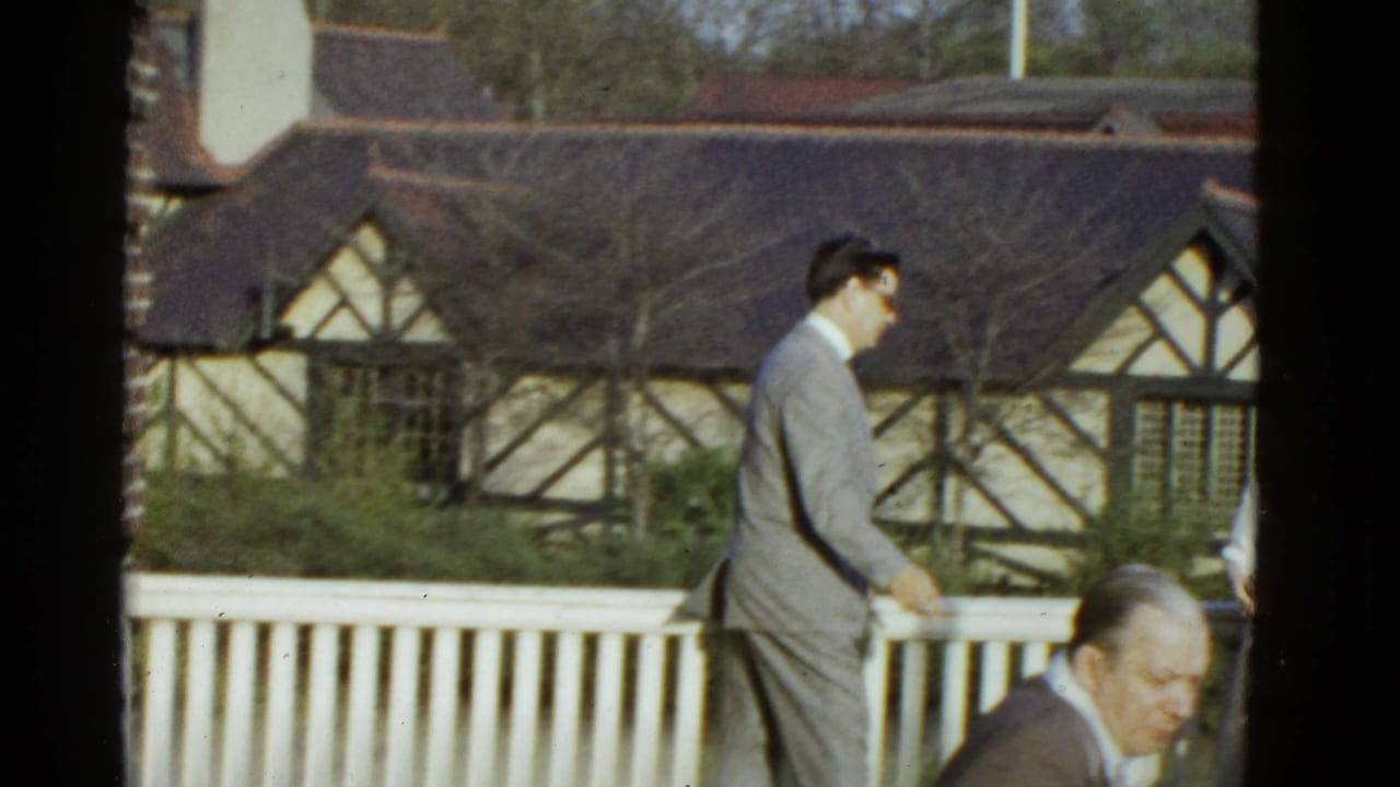 Man in a grey suit walks past a white fence with a Tudor-style building behind him.