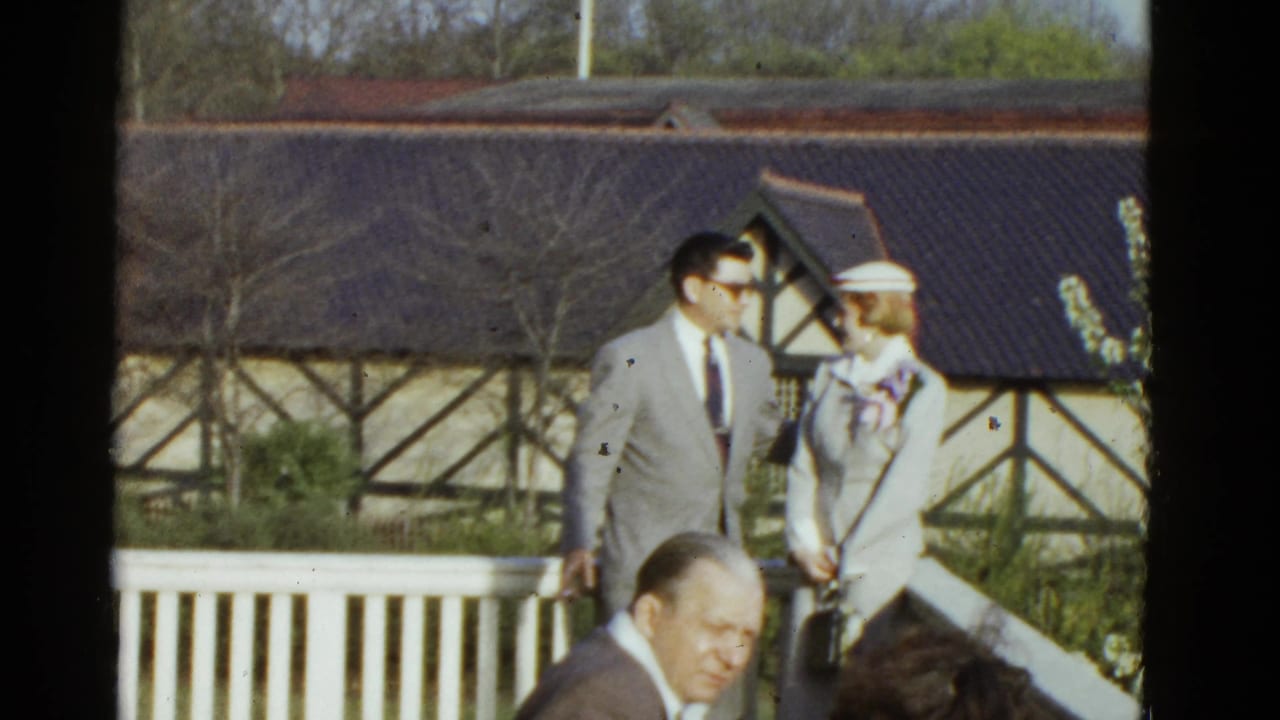 Man in grey suit and sunglasses stands near a woman in a white hat by a white fence.
