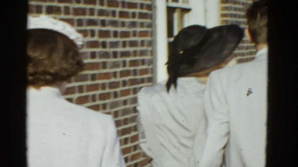 Adults in period coats and hats are filmed from behind near brickwork and a white railing during the same 1951 Boston outdoor scene.