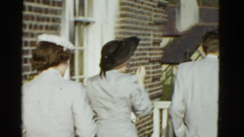 The camera turns toward nearby adults in coats and hats by brickwork, a window, and railing in the 1951 Boston home-movie setting.