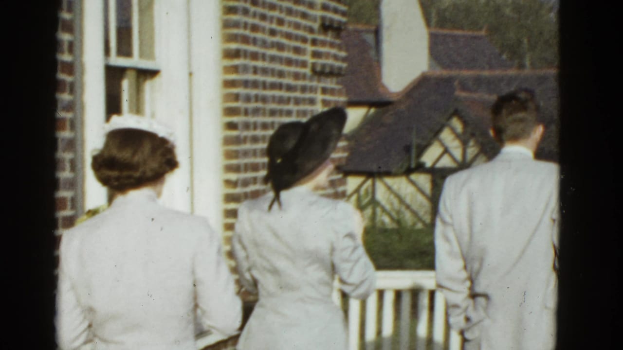 In 1951 Boston home-movie footage, two women in light suits and a man in a light suit stand on a porch facing away.