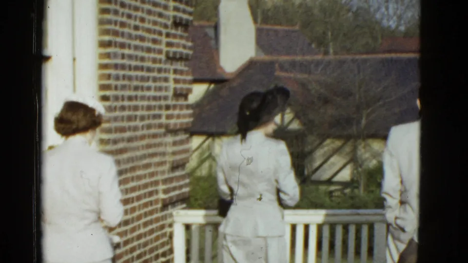 Two women in pale suits, one with a black hat, stand near a porch railing in the 1951 Boston exterior scene.