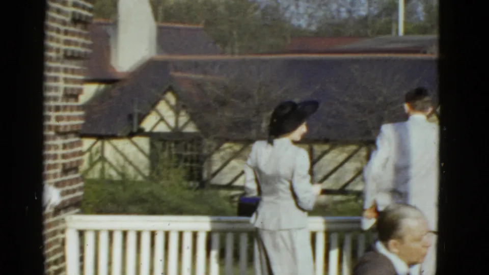 A woman in a black hat and pale suit stands near a man in a light suit on a porch in 1951 Boston footage.