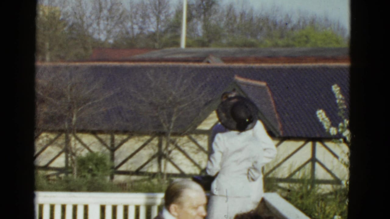 1951 Boston home-movie frame showing a woman in a light outfit and dark hat on a balcony, with a timber-framed building behind her.