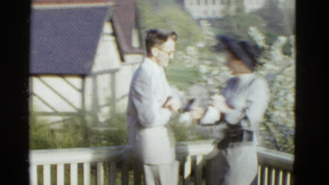 Man in a light suit leans toward a woman in a dark hat beside a balcony railing.