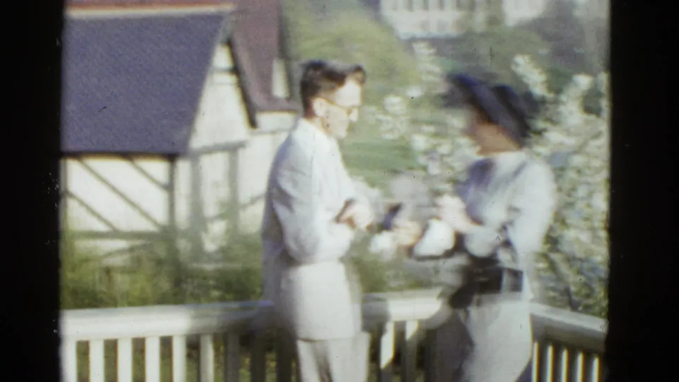 1951 Boston home-movie frame of a formally dressed couple sharing a close moment on a balcony with rooftops and flowers behind them.