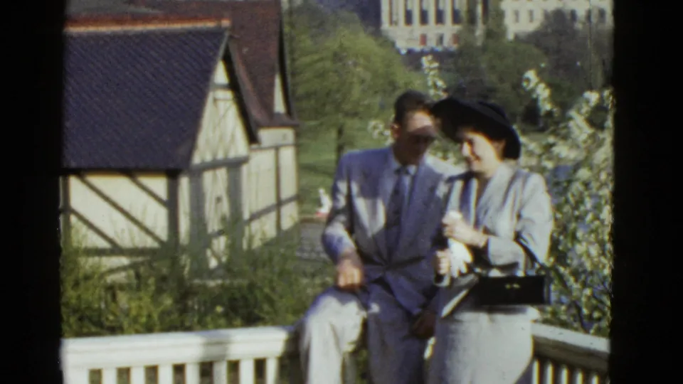1951 Boston home-movie frame showing a seated couple in formal clothing beside a white railing and flowering shrub.