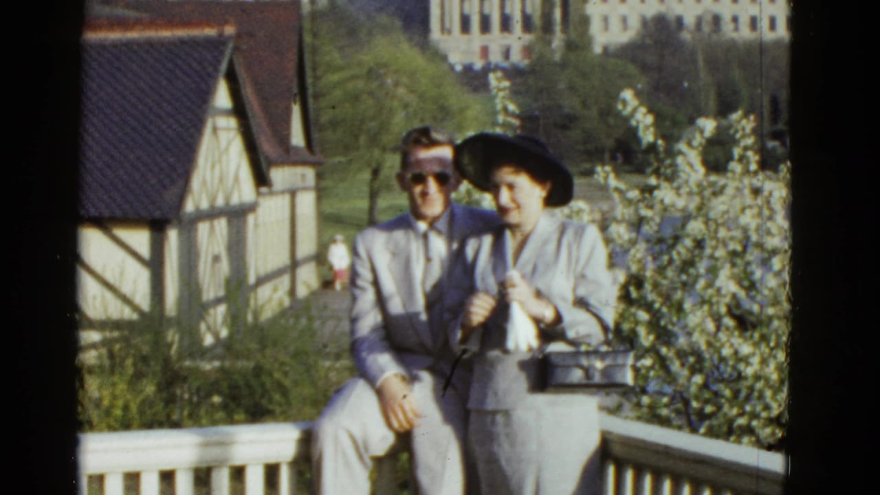 A well-dressed adult couple sits on a white railing in Boston, with flowering shrubs and a half-timbered building behind them.
