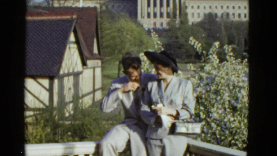 The couple remains seated outdoors in Boston as the man looks down and the woman turns slightly beside flowering shrubs.