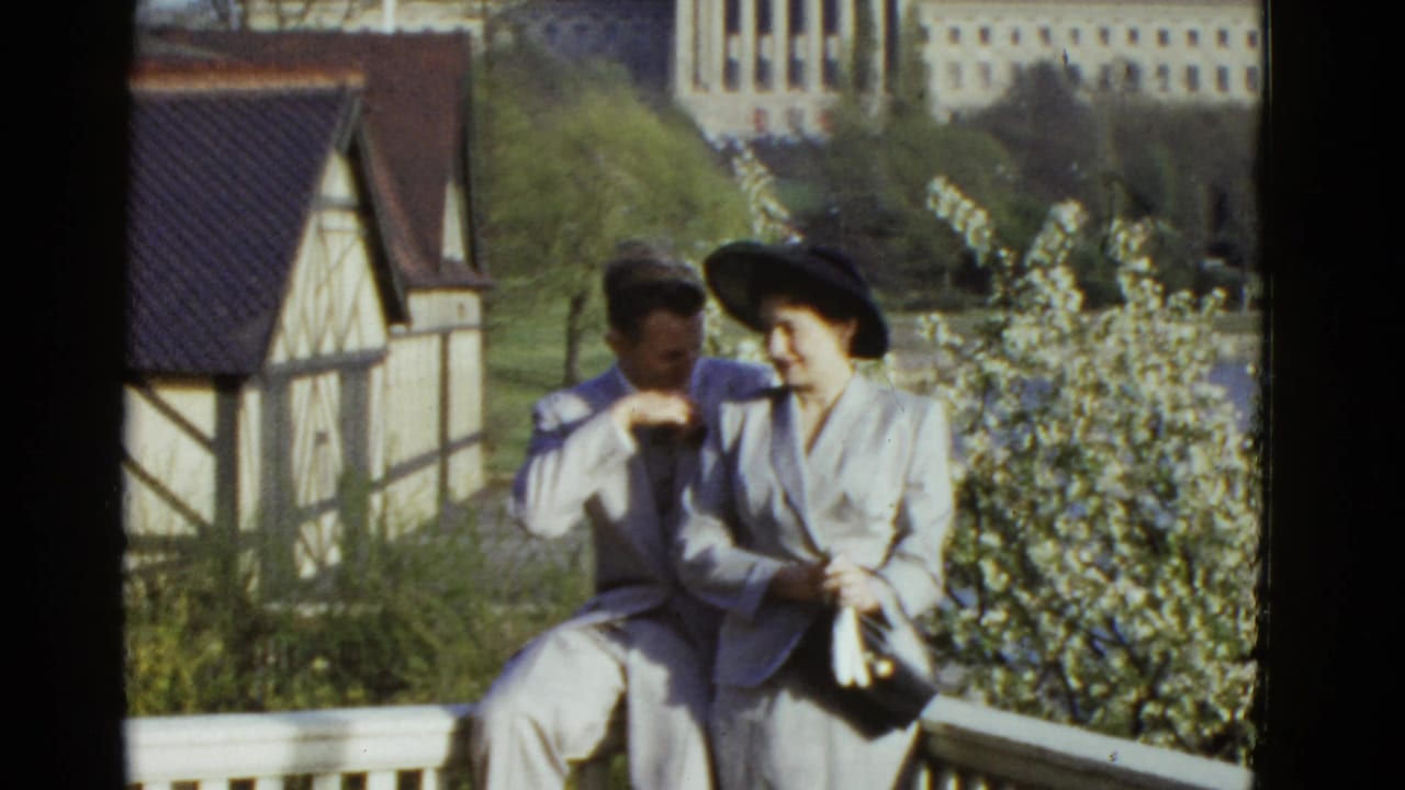 A man and woman pose on a white deck near flowering trees and a Tudor-style boathouse.