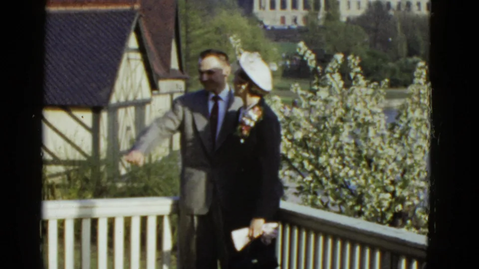 The formally dressed pair pauses on the deck, with the man gesturing and the woman holding accessories near the lakeside railing.