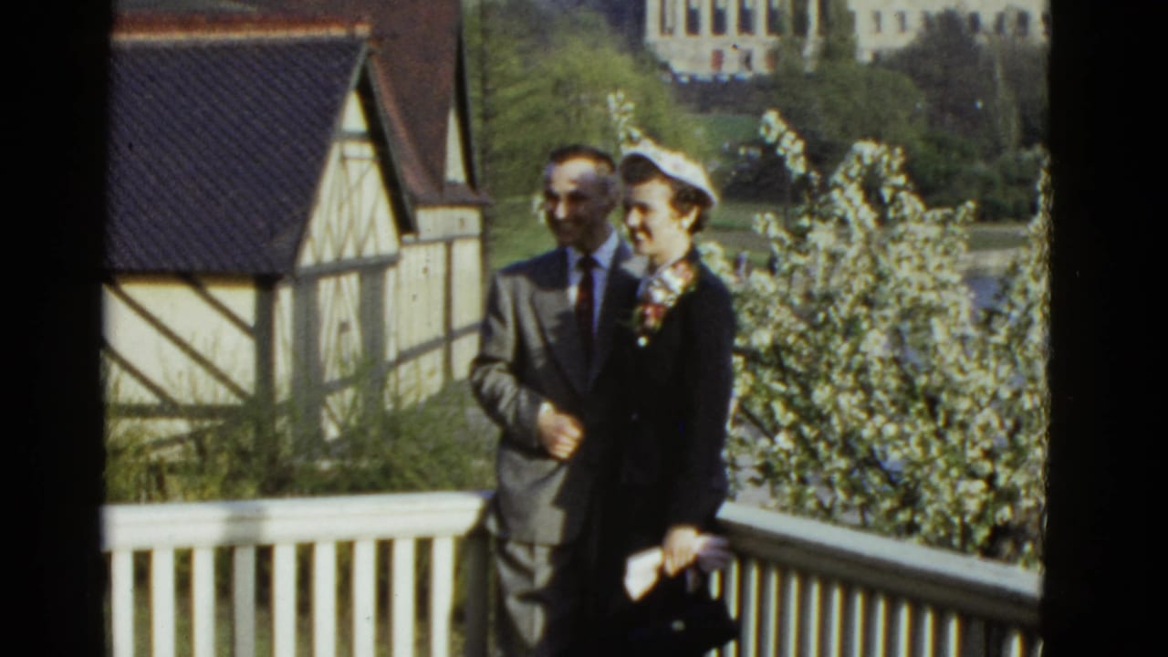 In 1951 Boston home-movie footage, a man in a gray suit and a woman in a dark coat with a white hat pose on a balcony.